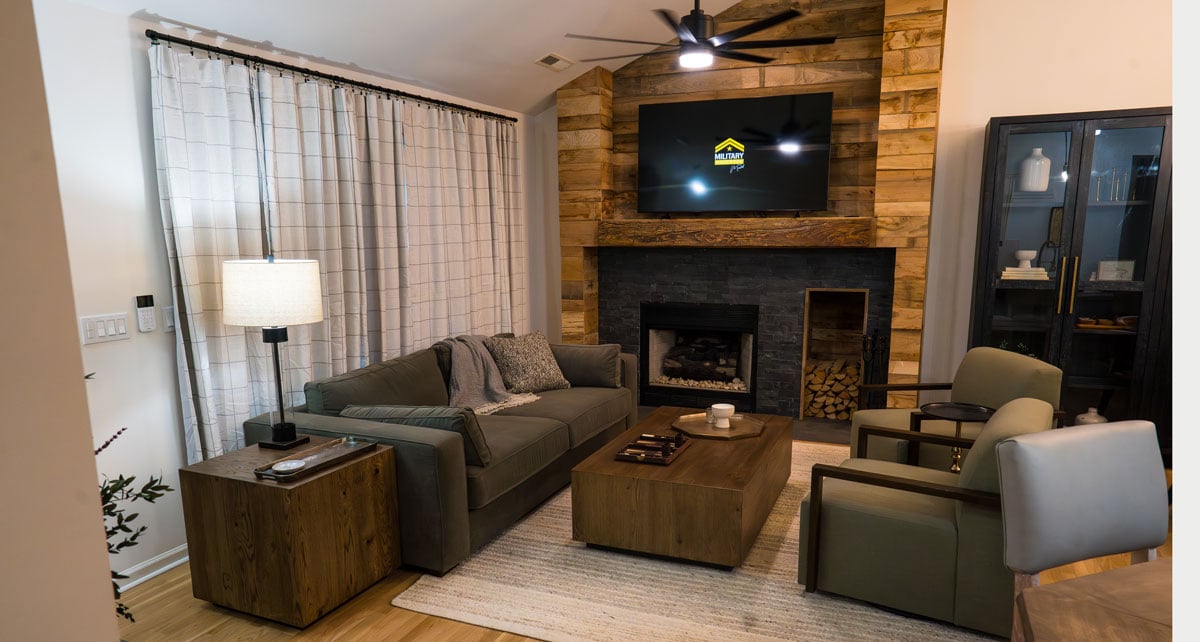 Modern living room with a gray sofa, two upholstered armchairs, and a wood coffee table facing a stone fireplace with a reclaimed wood accent wall. A wall-mounted TV hangs above the fireplace, with floor-to-ceiling curtains, a table lamp, hardwood floors, and a black display cabinet completing the warm, contemporary space.